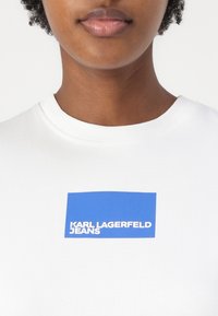 Biela bavlnená tričko s modrým obdĺžnikovým logom s nápisom "KARL LAGERFELD JEANS" v bielom písme, okrúhlym výstrihom a klasickým strihom.