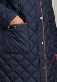 Μπουφάν σε σκούρο μπλε ναυτικό χρώμα με διαγώνιο σχέδιο, καφέ περιθώριο και μπροστινή τσέπη με κούμπωμα τρουκς. Μαλακή, επενδυμένη υφή.