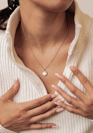 Woman wearing a smiley face pendant necklace, adjusting the collar of a white knit sweater, showing manicured nails with French tips.