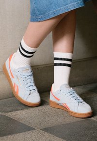 Sneaker in suede blu chiaro con accenti arancioni, suole in gomma marrone e lacci bianchi. Abbinati a calzini bianchi con strisce nere.