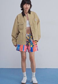 Khaki kurtka zapinana na guziki z brązowym kołnierzem, biała bluzka crop top oraz kolorowe szorty w paski z kwiatowym wzorem. Białe skarpetki i buty dopełniają strój.