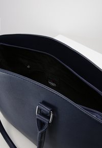 Námořnická modrá kožená taška s hladkou texturou, která má zipové uzavírání a vnitřní kapsu. Popruh obsahuje kovový prvek.