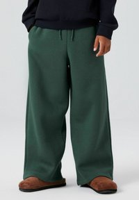 Bredde ben, mørkegrønne sweatpants lavet af blødt stof, med elastisk talje og snoretræk. Parret med brune, slip-on fodtøj.