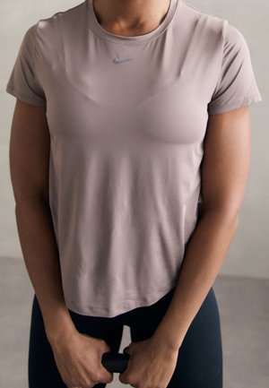 Personne portant un t-shirt Nike mauve clair tenant une barre d'exercice noire à deux mains, debout devant un fond uni.
