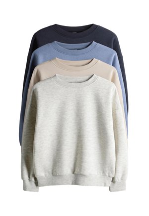 Kind met krullend haar dat een lichtgrijze trui draagt, wijde blauwe jeans en witte sneakers met zwarte strepen, staat met de handen in de zakken.