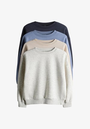 Kind met krullend haar dat een lichtgrijze trui draagt, wijde blauwe jeans en witte sneakers met zwarte strepen, staat met de handen in de zakken.