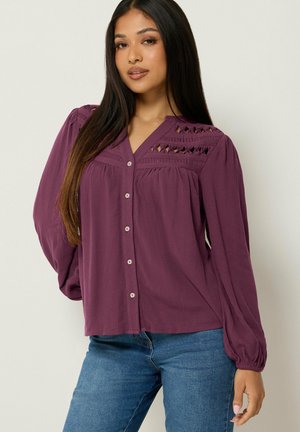 Frau mit langen dunklen Haaren trägt eine lila, langärmelige Bluse mit Cut-out-Details auf der Brust und blaue Jeans, steht vor einem hellen Hintergrund.