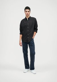 Camisa de botão xadrez preta com mangas enroladas, combinada com calças de veludo azul-marinho e ténis brancos, de pé contra um fundo simples.