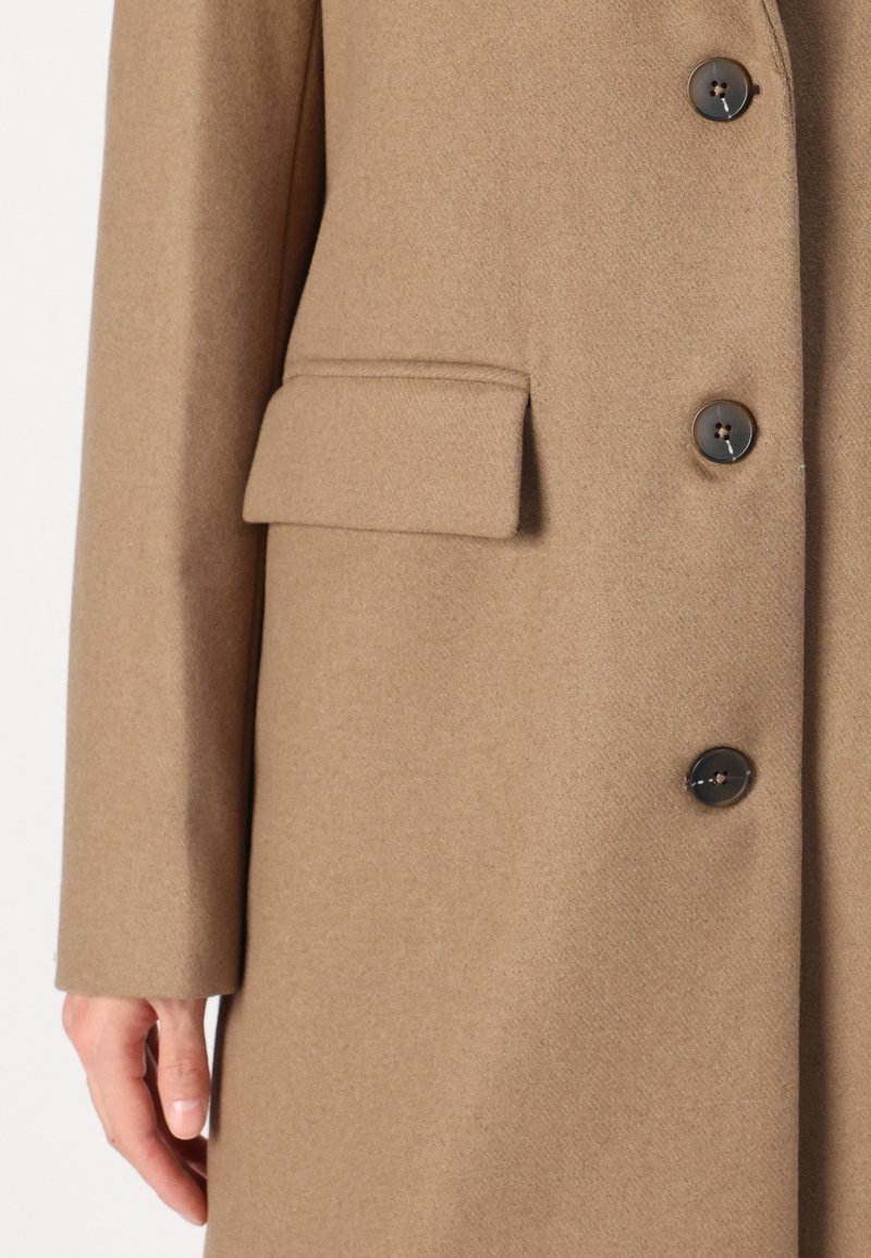 Cappotto di lana beige con tasca quadrata, bottoni neri e finitura testurizzata. Il design presenta una vestibilità su misura e linee pulite.