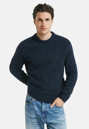 Jonge man met zwart haar, gekleed in een marineblauwe spikkelige trui en vervaagde blauwe spijkerbroek, staat met één hand in zijn zak tegen een witte achtergrond.