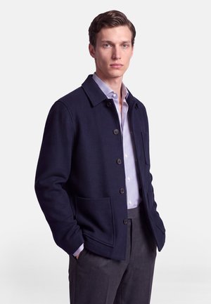 Homme portant une veste boutonnée bleu marine foncé sur une chemise blanche à rayures bleues et un pantalon gris foncé, debout les mains dans les poches.