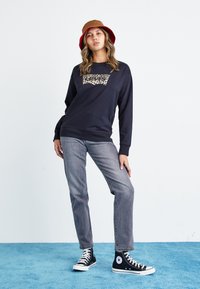Mornarska dukserica s printom leoparda na Levi's logo, sivi traperice i crne visoke patike. Model nosi smešnu kape u smešnoj i crvenoj boji.