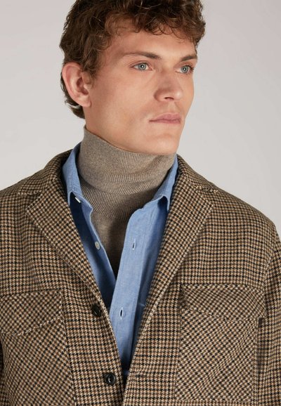Blazer a motivo houndstooth marrone con due tasche frontali, indossato sopra una camicia azzurro chiaro con bottoni e un dolcevita beige.