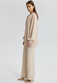 Blusa larga de textura beige con capucha, combinada con pantalones anchos beige y tacones puntiagudos beige. Tejido suave, diseño fluido y pliegues sutiles.