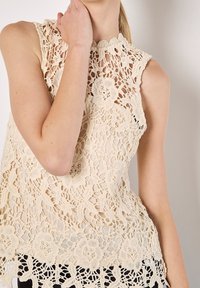 Haut en dentelle beige avec motifs floraux, col montant et design sans manches. Présente des détails complexes en crochet et un ourlet festonné.
