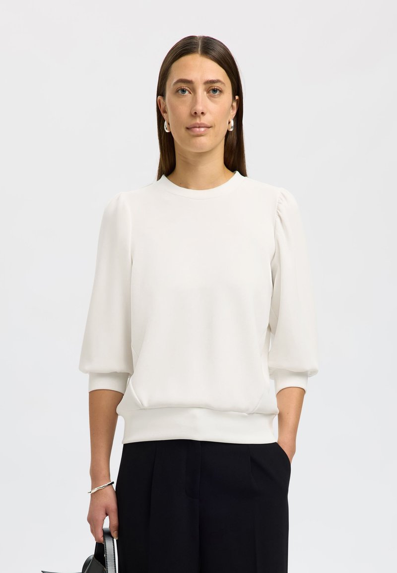 Weißer Pullover mit kurzen, gepufften Ärmeln, rundem Ausschnitt und lockerer Passform. Glatte Textur, einfarbig, kombiniert mit dunklen Hosen.