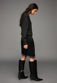 Šedý pletený sveter, čierna saténová sukňa s čipkovou ozdobou a čierne kožené čižmy po kolená s ostro špicatou špičkou. Bočný profil na neutrálnej pozadí.
