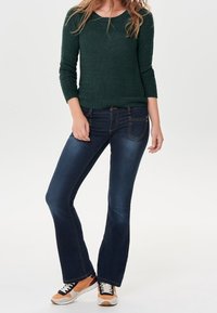 Jean évasé bleu foncé avec une taille ajustée, associé à un pull vert texturé à manches trois-quarts. Modèle portant des baskets orange et noires.
