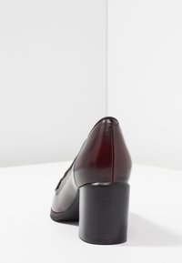 Chaussure à talon haut en cuir bordeaux avec un bout arrondi et un talon noir texturé ; surface lisse avec un minimum de détails de couture.