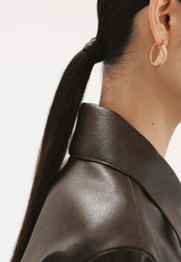 Pendiente de aro dorado con superficie texturizada, llevado en la oreja de un modelo, combinado con una chaqueta de cuero marrón con cuello alzado.