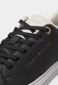 Μαύρο δερμάτινο αθλητικό παπούτσι με μαύρα κορδόνια, λευκή γλώσσα, χρυσό μεταλλικό κρικάκι και χρυσή ένδειξη "Tommy Hilfiger" στο πλάι.