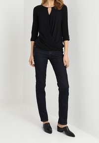 Top negro con detalle de ojal, mangas tres cuartos. Combinado con jeans de mezclilla oscuros y mules negros de punta afilada. Diseño simple.