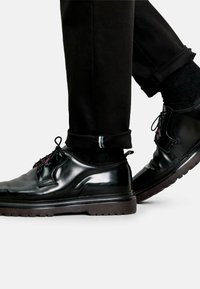 Zwarte leren schoenen met een gladde afwerking, ronde neus, veters en verhoogde rubberen zool. De broek is zwart en iets opgerold bij de mouwen.