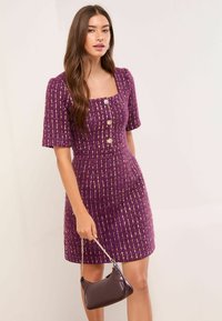 Rochie mov din tweed cu mâneci scurte, decolteu pătrat, trei nasturi în față și siluetă mulată. Modelul ține o geantă mică, închisă la culoare.