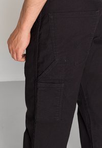 Närbild av en persons hand bredvid svarta cargobyxor med sid- och bakfickor, synliga sömnadsdetaljer på tyget.