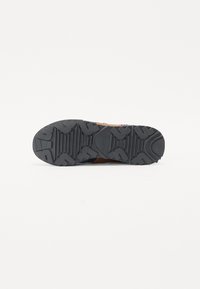 Semelle de chaussure de sport présentant un motif en caoutchouc noir texturé avec des rainures profondes pour l'adhérence, contrastant avec une partie supérieure marron.
