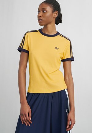 Kobieta w żółtej koszulce Adidas z granatowymi lamówkami i paskami, zestawiona z granatową spódnicą z białymi paskami.