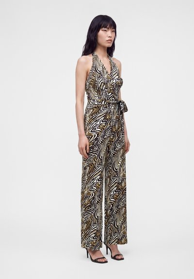 Femme portant une combinaison sans manches à col halter avec un motif abstrait tourbillonnant noir, blanc et doré, associée à des sandales à talons hauts noires.