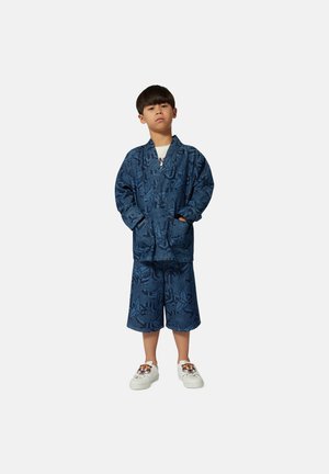 Blauwe, gemêleerde oversized jas gecombineerd met bijpassende shorts, beide met abstracte ontwerpen. Witte sneakers met een tijgerafbeelding.
