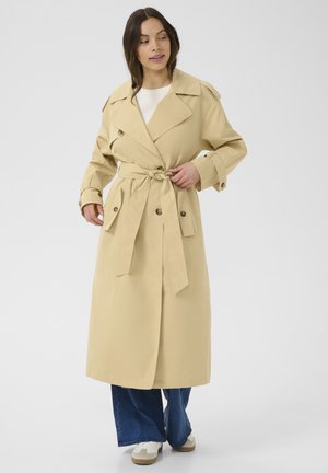 Beige színű, övvel ellátott trench coat, nagyméretű hajtókákkal, dupla gombos záródással és oldalsó zsebekkel. Kék, széles szárú farmernadrág felett viselve.