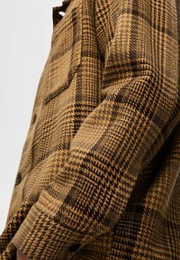 Brązowa i musztardowa koszula w kratę, wykonana z teksturowanej tkaniny, z zapięciem na guziki z przodu i jedną kieszenią na piersi. Udekorowana różnymi wzorami.