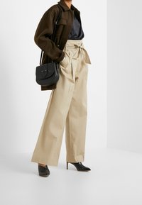 Person som bär beige högmidjade vida byxor med en knyten bälte, svart silketopp, mörkbrun jacka, svarta klackskor, och bär en svart axelremsväska.