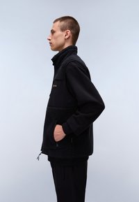 Black fleece vest with a high collar, zippered pockets, and a smooth fabric overlay. The design features contrasting textures.