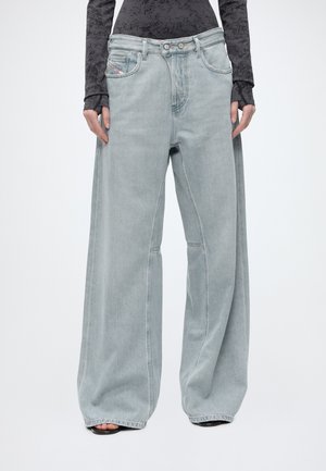 Osoba nosząca bardzo szerokie jasnoniebieskie dżinsy denim i długi czarny top w wzory, ręce luźno opuszczone wzdłuż ciała, stopy w sandałach z odkrytymi palcami.