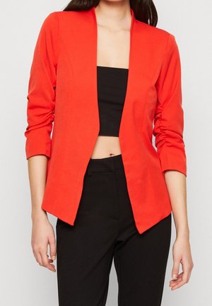 Femme portant un blazer ouvert rouge vif sur un top court noir et un pantalon noir taille haute, sur un fond clair uni.