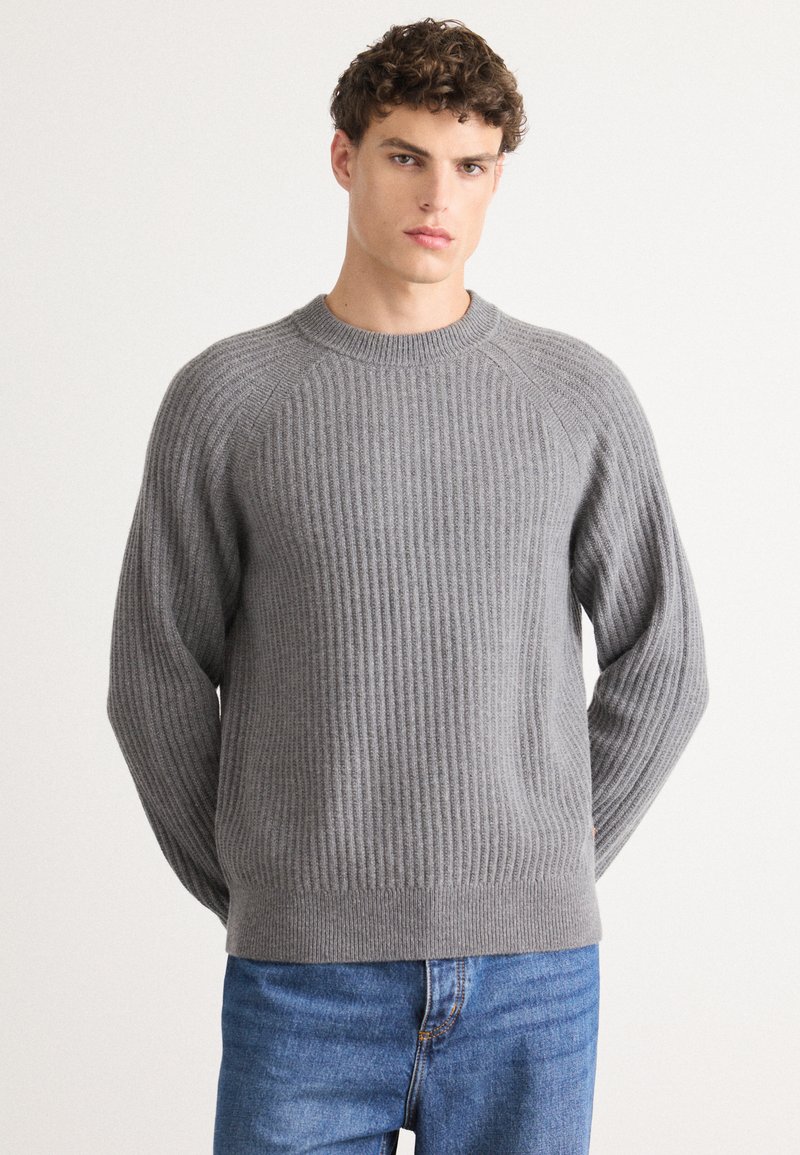 Grauer, gerippter Pullover mit Rundhalsausschnitt und Raglanärmeln, aus strukturiertem Stoff hergestellt; kombiniert mit blauen Jeans.