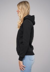 Black hoodie with a front pocket, ribbed cuffs, and a hood, displayed on a person wearing light blue jeans, viewed from the side.