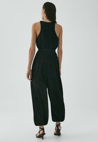 Mustassa haalariasussa on olkaimeton muotoilu, vyötärö on kiristetty, ja lahkeet ovat leveät. Materiaali on sileä ja kangas virtaa kauniisti. Takana on korkea kaulus.