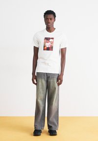 Λευκό μπλουζάκι κοντομάνικο με κεντρική φωτογραφική εκτύπωση, συνδυασμένο με χαλαρού τύπου τζιν σε ξεθωριασμένο γκρι χρώμα και μαύρα αθλητικά παπούτσια.