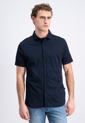 Ung mand med krøllet hår iført en mørkeblå kortærmet skjorte med knapper og lyseblå jeans, stående mod en enkelt hvid baggrund.