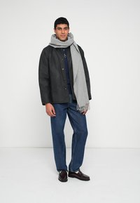Black waterproof jacket, gray knitted scarf, dark shirt, blue denim jeans, brown shoes, standing against a white background.