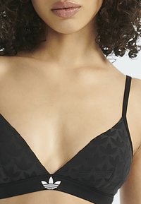 Kobieta nosząca czarny trójkątny biustonosz z subtelnym wzorem i logo Adidasa umieszczonym na środku pasa, ukazująca ramiona i górę klatki piersiowej.