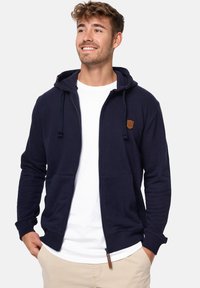 Tamno plava zip-up dukserica s vezicama, prednjim dzepovima i smeškanom kožnom oznakom. Napravljena od mekanog, teksturiranog materijala sa rebrastim manžetama.