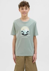 Vaaleanvihreä puuvillainen T-paita, jossa on pyöreä graafinen kuva auringonlaskusta, palmuista ja teksti "Jack & Jones Worldwide" mustalla värillä.