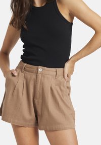 Ruskean pellavasekoitteen shortsit, joissa on pleated etuosa, vyötärön nappikiinnitys ja sivutaskut, yhdistettynä mustaan hihattomaan toppiin.