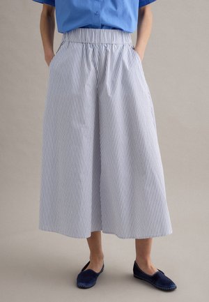 Persona che indossa pantaloni bianchi larghi e ampi con sottili righe verticali, abbinati a scarpe slip-on blu scuro e una camicia blu.